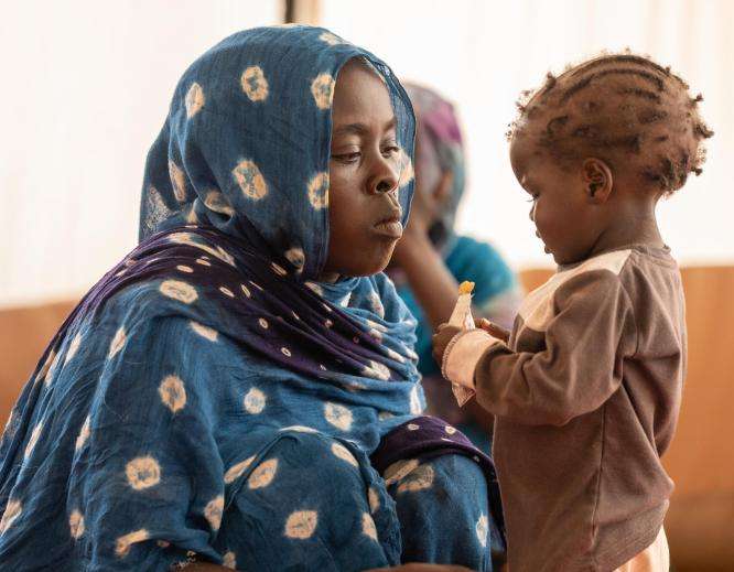 Mother in a blue hijab watches her child eat therapeutic peanut paste in Adré, Chad.