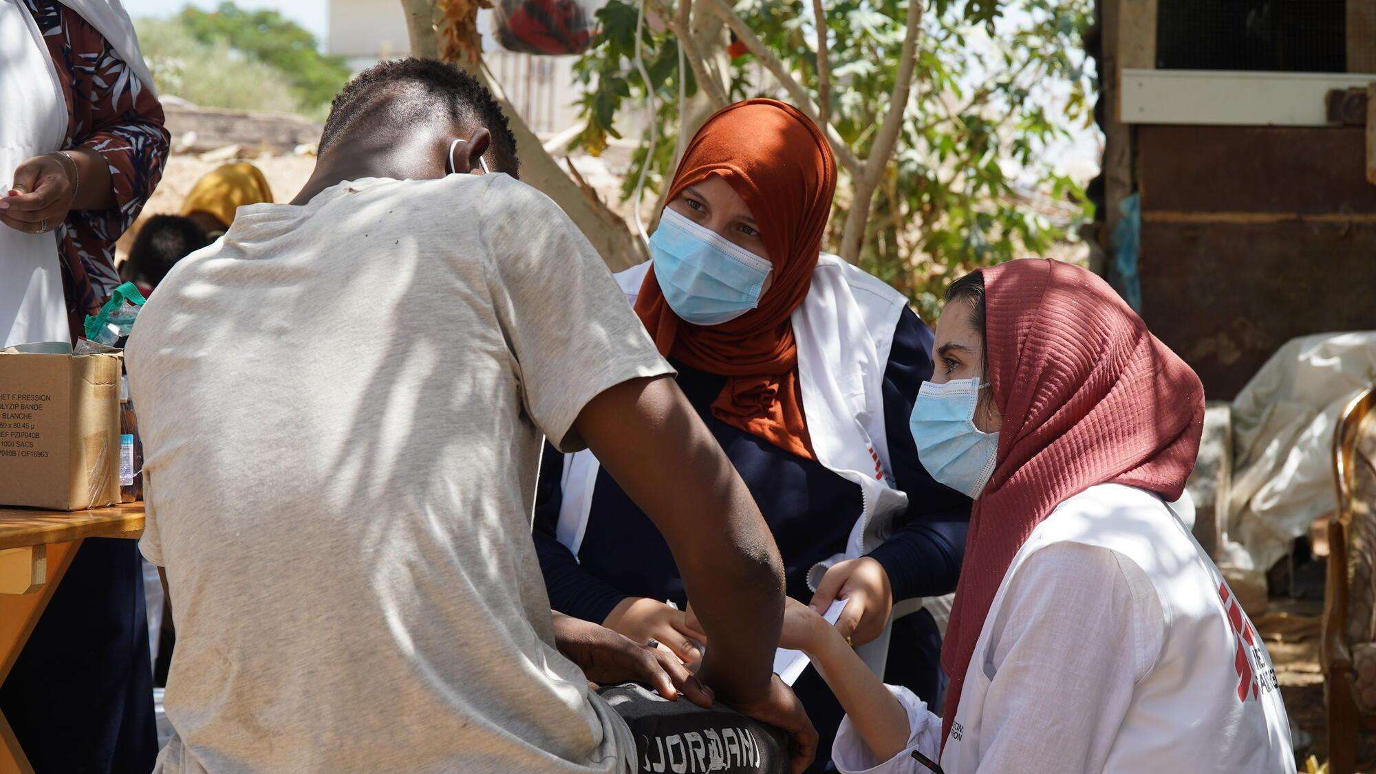 MSF staff members wearing face masks, headscarves, and white vests speak to a man with back turned in Libya. 