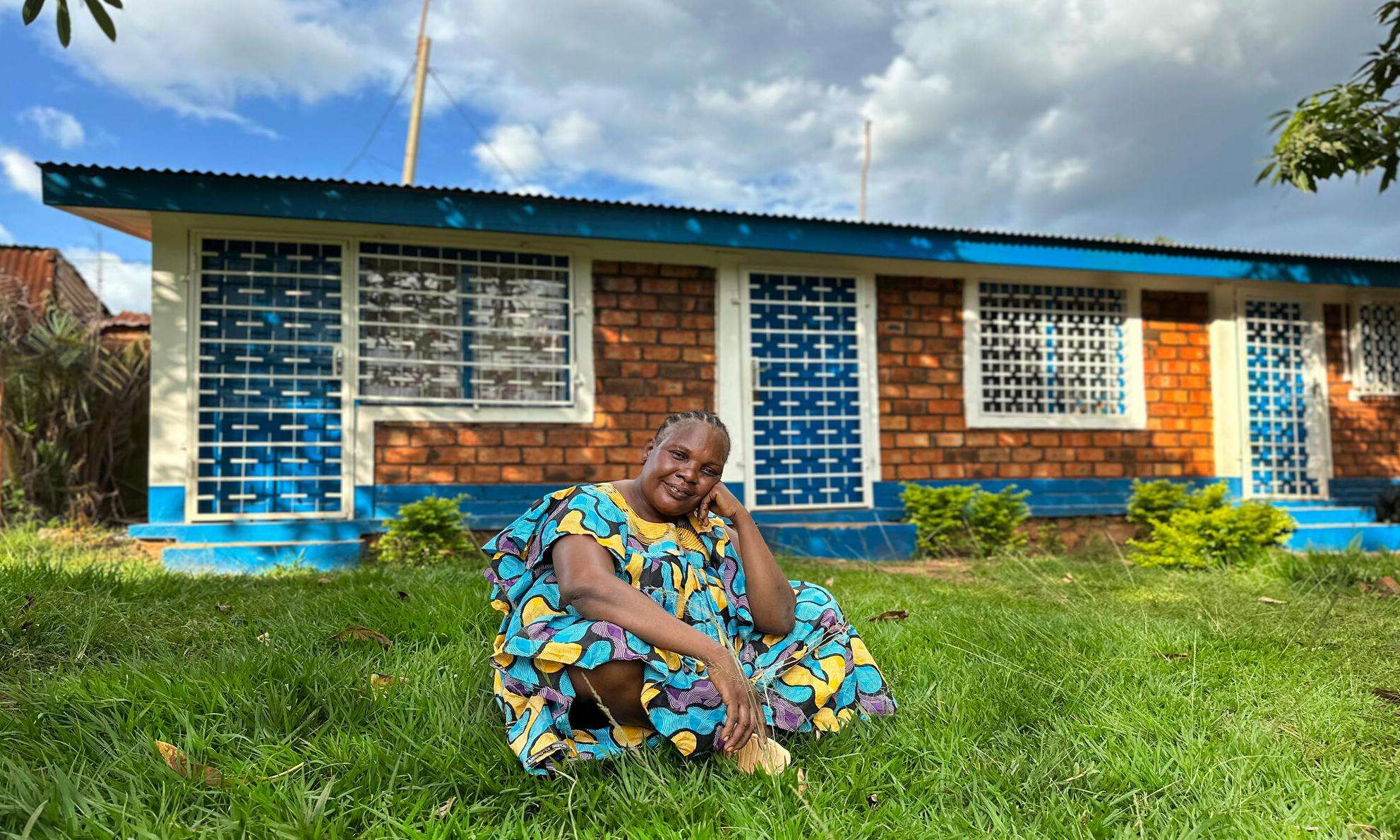 A smiling woman in brightly colored dress sits on the lawn in front of a brick building in Central African Republic.