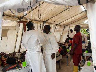 Two medics in white robes are facing each other adjusting their robes inside a white tent surrounded by patients. 