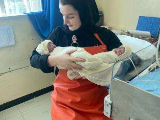 A woman in a head-covering and orange-ish uniform holds a small newborn wrapped in a blanket. 