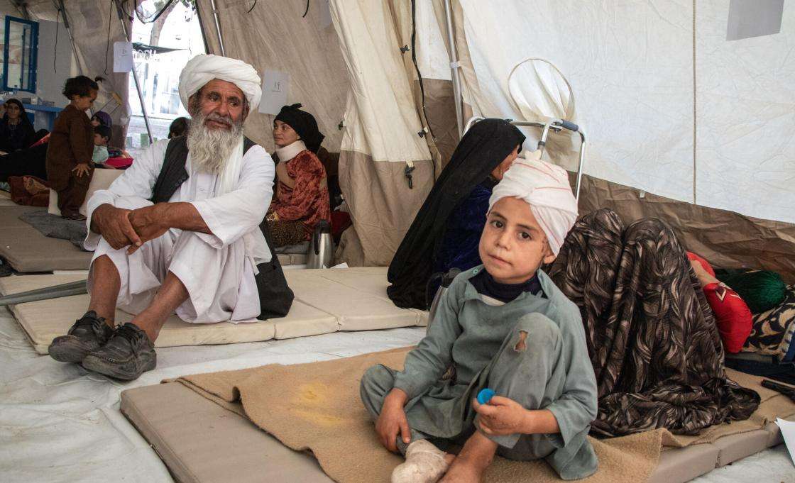 A young child with a bandage on his head sit on the floor of MSF's medical tent next to his grandfather