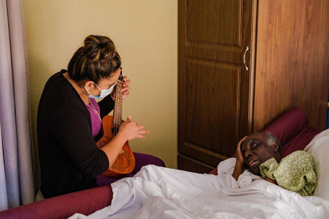 Masked woman sitting next to a patient's bedside playing the ukulele for patient.
