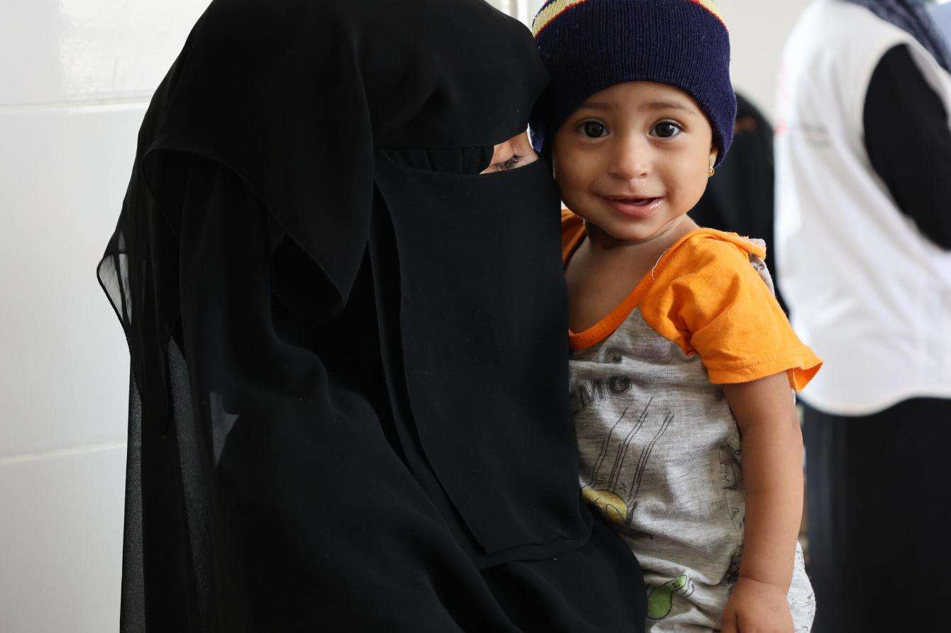 A woman in black niqab holds a child in yellow T-shirt at an MSF health facility in Yemen.