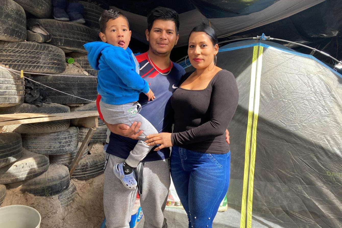 A migrant family including mother, father, and child stand together at a shelter in Mexico. 
