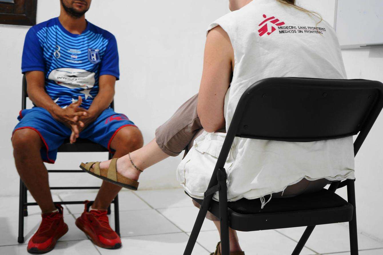 Two people whose faces we cannot see sit on black plastic chairs, facing each other. One is wearing a blue T-shirt the other is in an MSF vest.