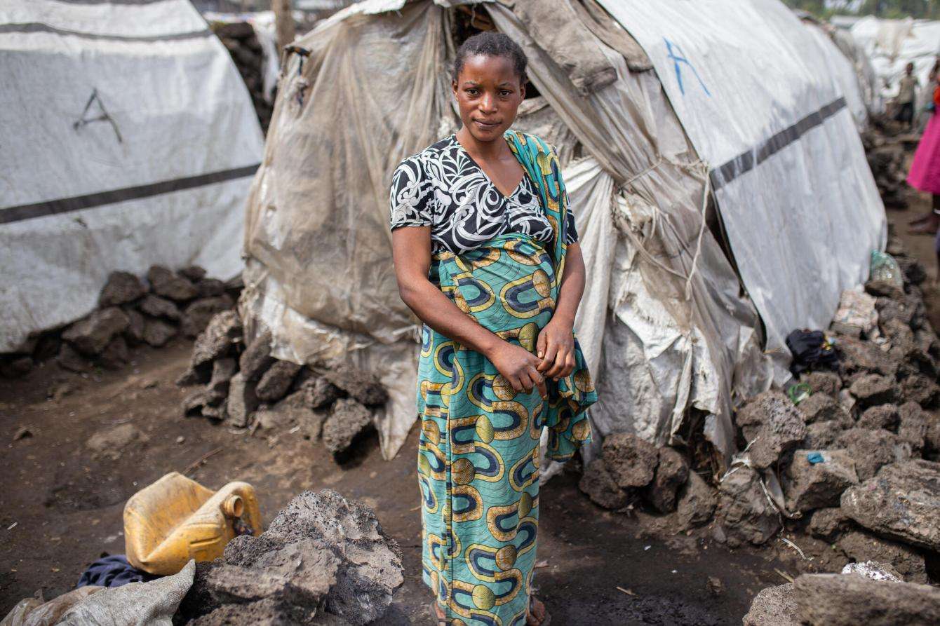 A woman in a green dress stands before makeshift tents in a displacement camp near Goma, Democratic Republic of Congo.