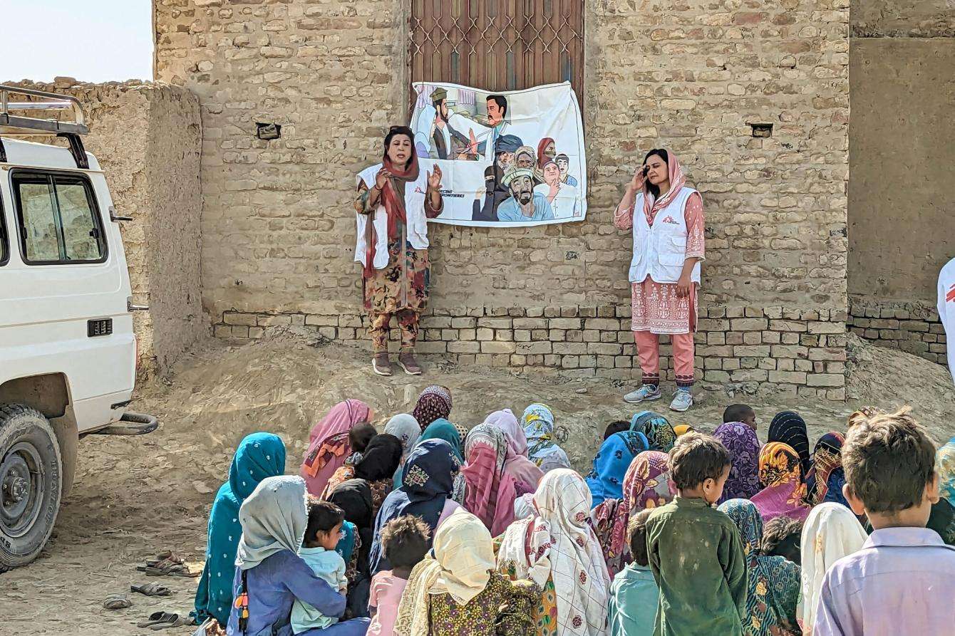 Two women in MSF vests stand outdoors with posters on the wall and an audience of people sitting in front of them in Dadu district, Pakistan.