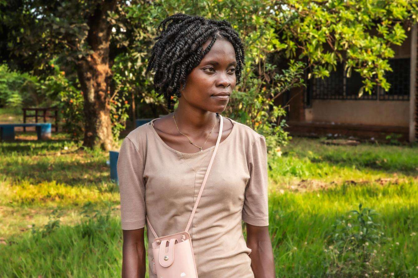 A young woman in beige T-shirt stands beside trees in a grassy area in Central African Republic.