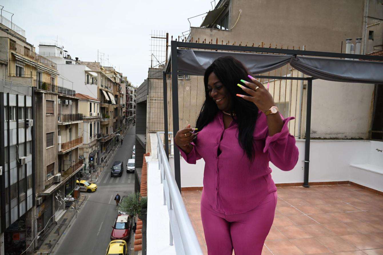 A trans woman asylum seeker in a pink outfit stands on a balcony in Greece looking down at the street.