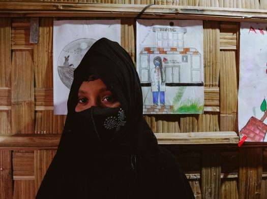 A Rohingya girl wearing a black veil standing beside drawings