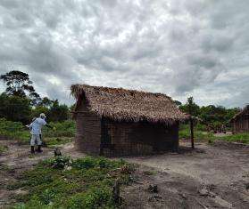 An MSF staff member is walking through a rural area to a hut with a thatched roof. 
